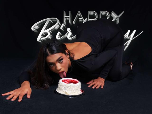 SamanthaStele, with black dress and long hair, kneels eating a white cake with red filling, tongue out, black background, shiny "Happy Birthday" letters behind her.