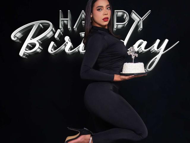 SamanthaStele with dark skin, red lipstick, black long-sleeve top, black pants, holding white cake, black heels, standing, holding cake, black background, "Happy Birthday" neon sign.