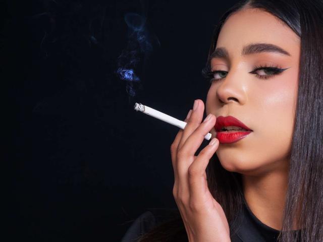 Samantha Stele, con rossetto rosso, capelli neri e lisci e occhi truccati in stile smokey, che porta una sigaretta accesa alle labbra, con il fumo blu che si alza..