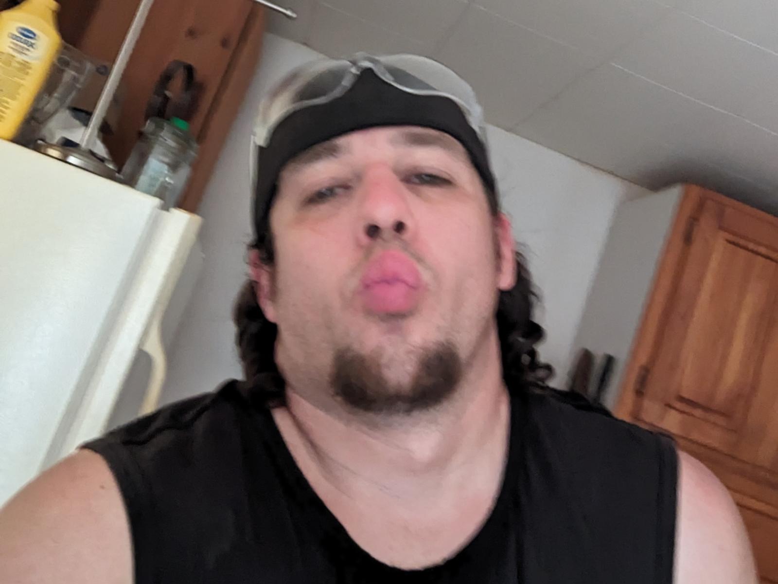 Nataurius69 with a black headband, goggles, and a goatee, puckering his lips in a goofy expression, wearing a black sleeveless shirt, in a slightly blurry photo.