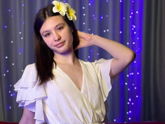 Harisma with a yellow plumeria flower in her brown hair, wearing a white ruffled top, has her hand behind her head, looking cute with small tits and a subtle smile.