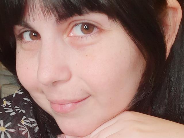IvyBloom's face, showing pale skin, brown eyes, black straight hair with bangs, slight smile, and pink lips, resting chin on hand, wearing a black floral shirt.