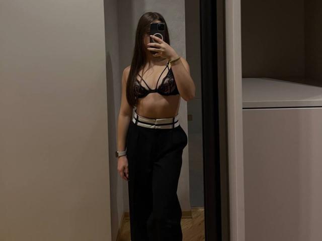 PaigePrado with long brown hair, wearing a black lace bra, showing small tits, black high-waisted pants, white belt, holding phone, mirror selfie, wooden floor, dim room.