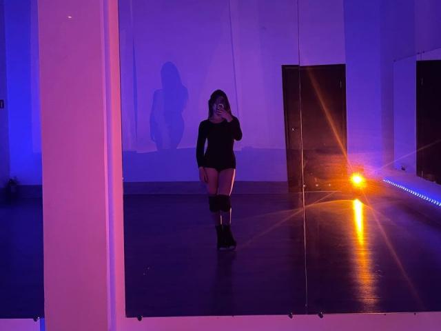 PaigePrado with dark hair, wearing black long-sleeve top and black shorts, holding phone, shadow on wall, purple-blue lighting, orange light on right, standing on dark floor.