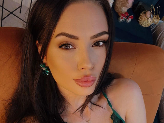 MonaHott69 tiene labios rosados y finos, cabello castaño oscuro, aretes verdes y ojos marrones. Está acostada en una silla de cuero marrón, mirando directamente a la cámara con una expresión seria..