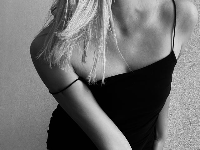 Black-and-white photo of PrimeS with blonde hair, wearing a spaghetti-strap black dress, showing a slinky, slim upper body and a single strap sliding off their shoulder.