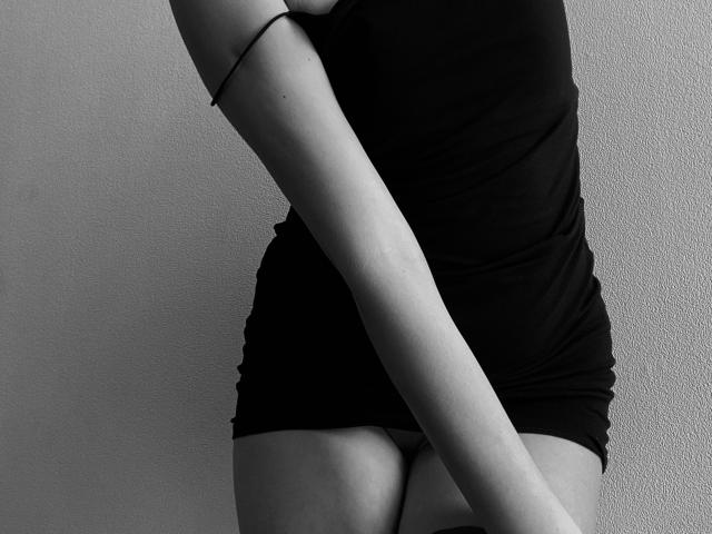 Black and white photo of PrimeS with a slim, toned body, wearing a tight black dress, arms crossed over the chest, showing a small butt and bare thighs.