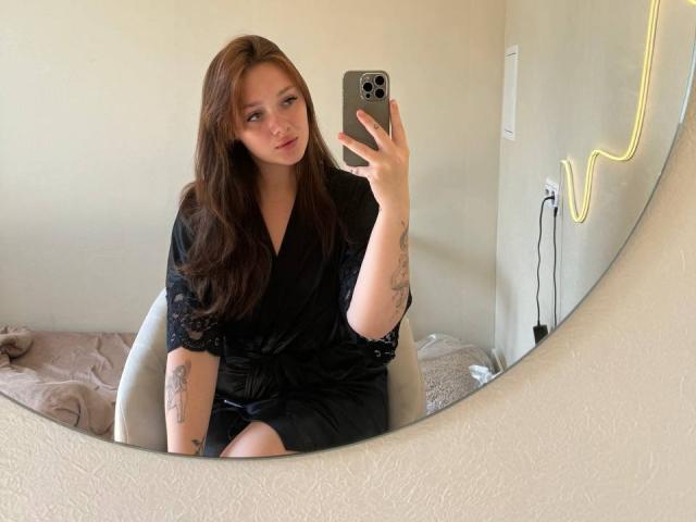PolishaMi, con cabello largo y castaño, vestido de encaje negro, tatuajes en los brazos, sosteniendo un teléfono, tomando una selfie en el espejo, sentada en una silla, con una sonrisa suave, piel clara, luz de neón en la pared, cama en el fondo..