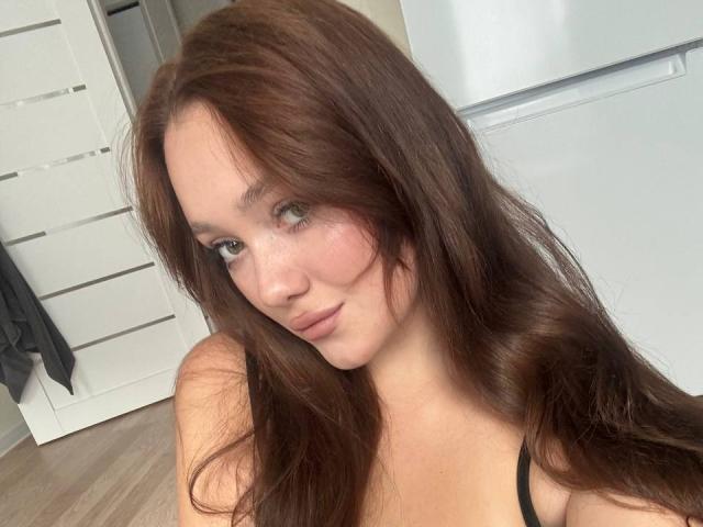 PolishaMi tiene el pelo largo y castaño, ojos verdes y una ligera sonrisa, lleva una blusa negra que deja ver sus pechos y tiene un rostro adorable con pecas, en un dormitorio..