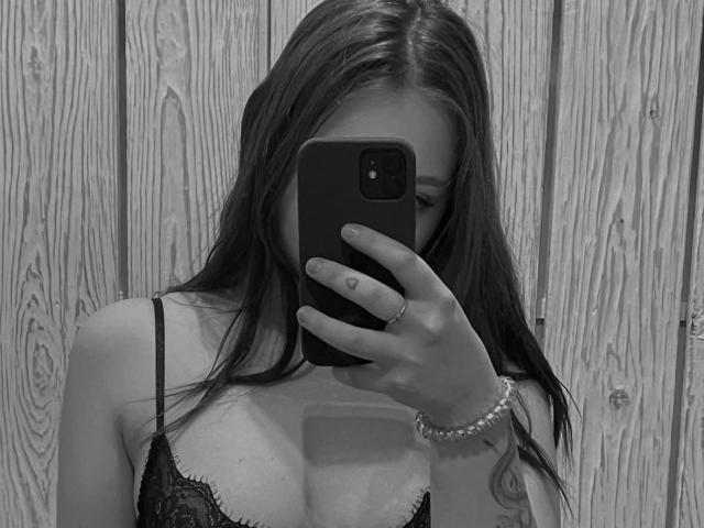 PolishaMi, con cabello largo y oscuro, vestida con un sujetador de encaje negro, tomando un selfie con un teléfono, cubriéndose la cara, mostrando un tatuaje en su mano derecha y una pulsera de perlas..