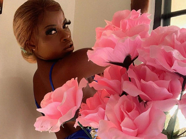 Essyreina69-hot with dark skin, blonde hair, and blue bra, looking over shoulder at pink roses, wearing green earrings, in a sunlit room.