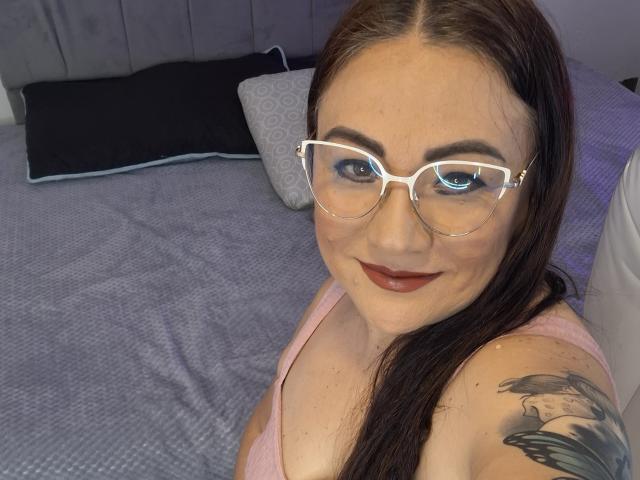 LadyNylon: Una donna con gli occhiali, rossetto rosso e capelli castano scuro, sorride alla fotocamera, mostrando un tatuaggio sulla spalla destra, indossa una maglietta rosa e siede su un letto..