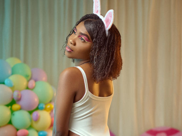 Alana Patel mit lockigem, braunem Haar, die ein weißes Tanktop trägt, pinken Lidschatten und pinke Hasenohren, blickt zurück und steht neben bunten Ballons. Ihr Hintern ist sichtbar..