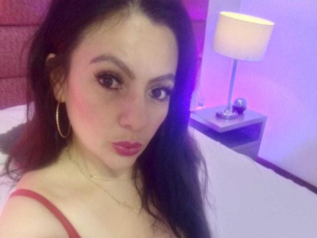 JakyKidman, con rossetto rosso, orecchini a cerchio e una collana d'oro, ha i capelli scuri, un leggero blush rosa e indossa una canottiera rossa che mette in mostra il suo seno in una camera da letto..