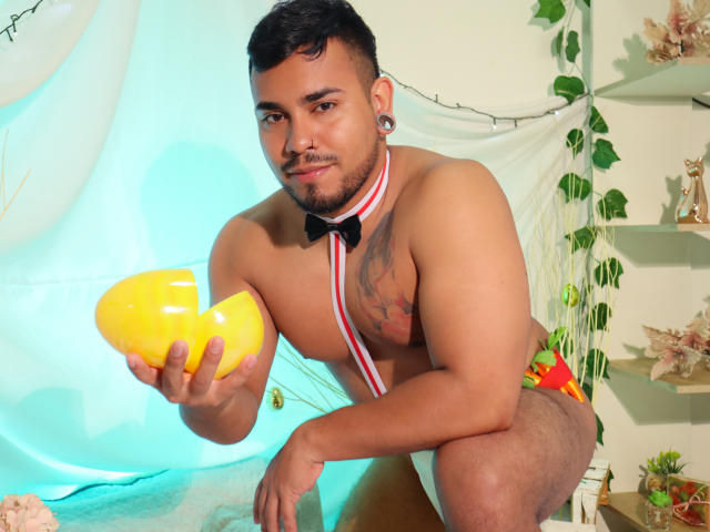 BrianMarly with a muscular build, dark hair, and a beard, holding a yellow melon, wearing a red and white striped bowtie, and colorful underwear, showing a tattoo on his chest.