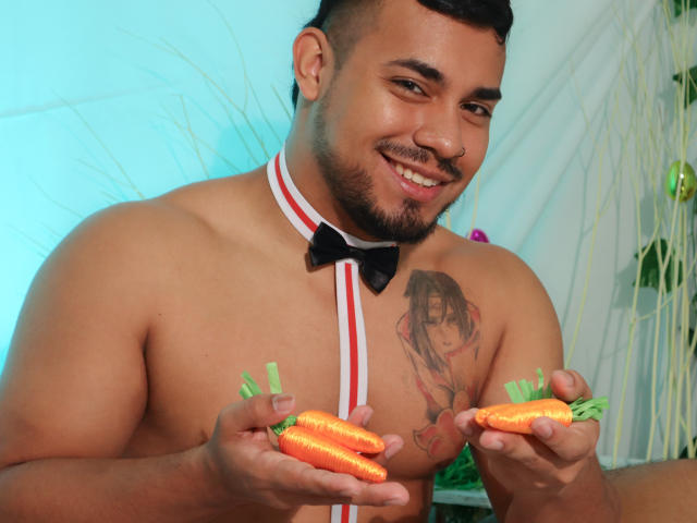 BrianMarly, shirtless with a black bowtie, holding two carrots, smiling at the camera. He has a tattoo of a woman on his right chest and a nose ring.