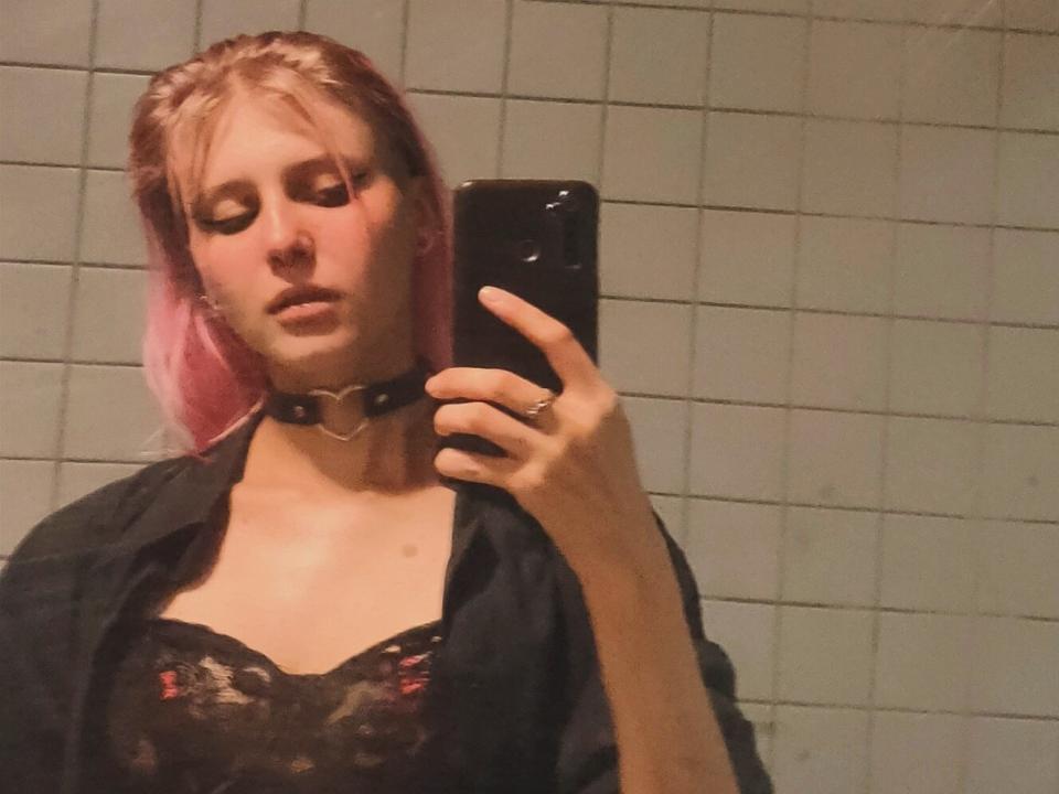 VelvyMonica with pink hair, black choker, and lace bra, holding phone in bathroom mirror, wearing open shirt, looking down, ring on finger, tits visible, medium breast size.