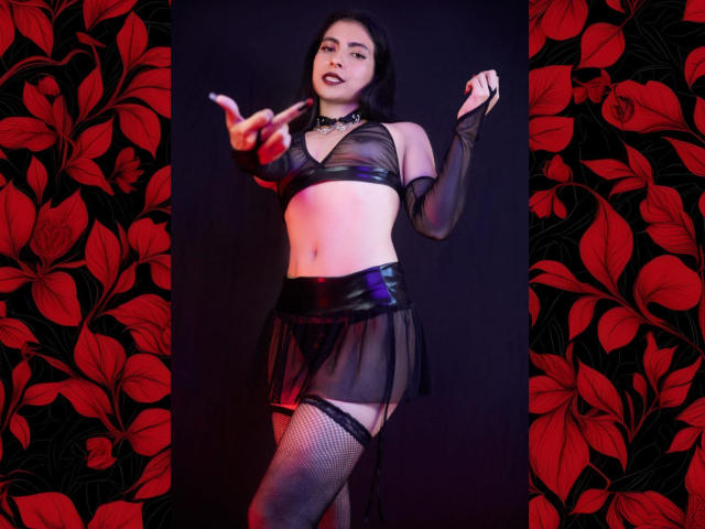 SamantaKi with dark hair, wearing a sheer black top and skirt, black thigh-highs, and fishnets, pointing with her right hand, red lips, choker, and a confident expression.