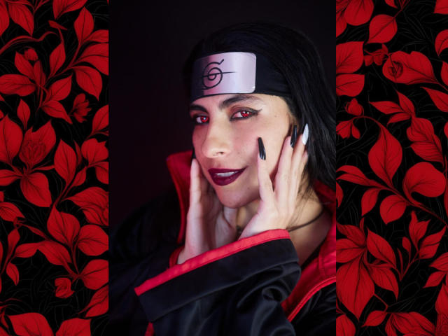 SamantaKi with black headband, red eyes, dark lipstick, black nails, hands on face, smiling, wearing black and red ninja outfit, floral red and black pattern on sides.
