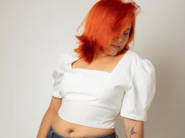 RubiQueenS mit leuchtend orangefarbenem Haar, die ein weißes Crop-Top mit Puffärmeln trägt, ein Tattoo auf ihrem rechten Arm zeigt und blaue Jeans trägt, leicht nach vorne gebeugt..