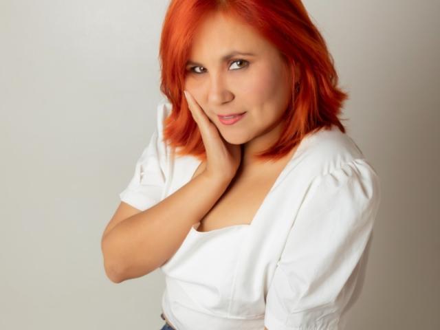 RubiQueenS with red hair, white shirt, hand on cheek, smiling, small tits, cute, light skin, soft makeup, casual pose, studio shot.