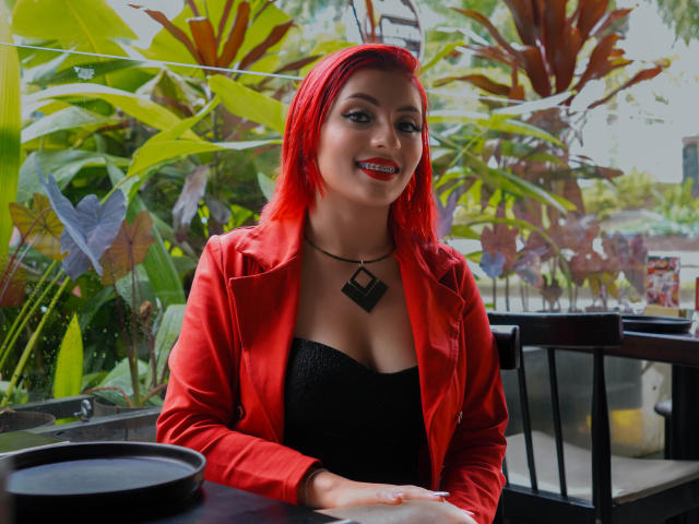 LizBonnet with bright red hair, red jacket, black top, and diamond necklace, smiling with red lipstick, sitting at a table with a black plate, hands on the table.