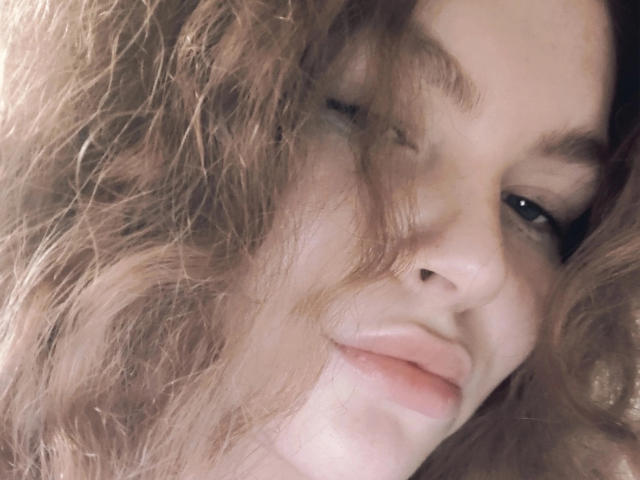 Mable Jocelyn tiene el cabello castaño ondulado que le cubre el rostro, con labios rosados ligeramente separados y una nariz pequeña, y mira hacia abajo con una sonrisa sutil y segura..