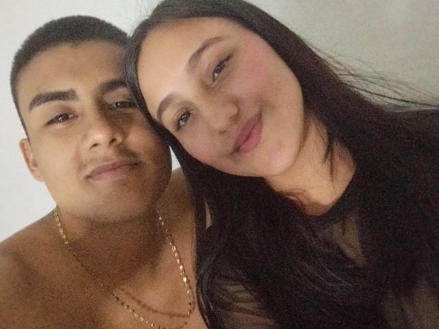 MelissaAndJuan and Juan, shirtless with a gold chain, next to Melissa with long black hair, smiling, and leaning in. Her tits are slightly visible through a sheer top.