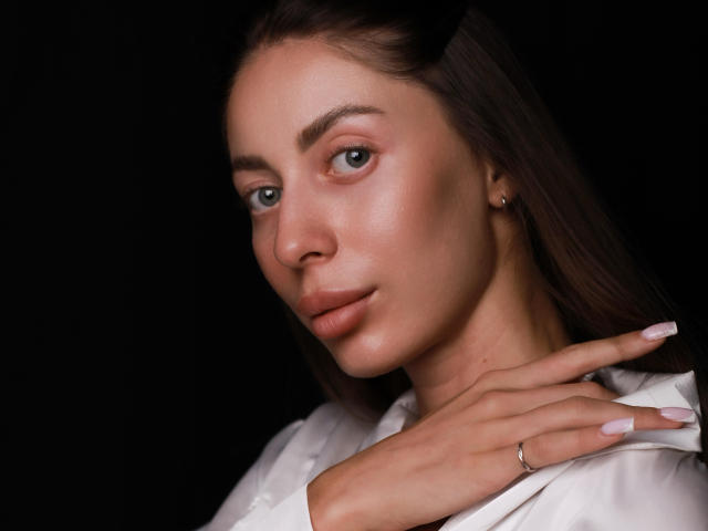 HotBabeArt has light skin, blue eyes, and brown hair, with a slight smile, wearing a white shirt, touching her neck, showing a ring, and having pink nails.