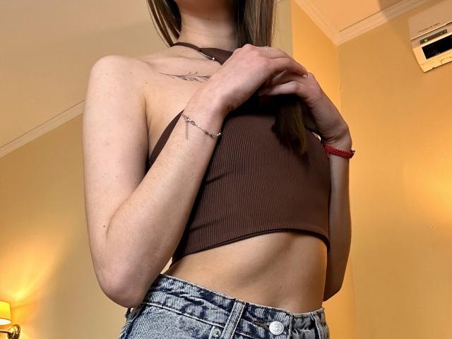 Karish wearing a brown ribbed crop top, showing her small tits and flat ass, with denim jeans, a red bracelet, and a tattoo on her left wrist.