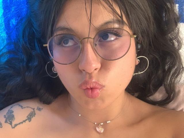 MadyJackson with puffy lips, wearing round glasses, hoop earrings, and a heart tattoo on her shoulder, has a necklace with a pink heart. She has dark, wavy hair.