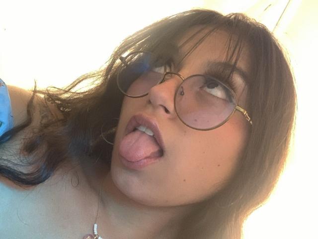 MadyJackson with round glasses, brown wavy hair, and a nose piercing, sticking her tongue out, showing off her small, cute ass and a subtle lip piercing.