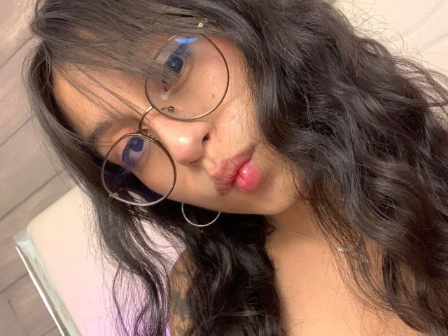 MadyJackson with long, wavy black hair, big round glasses, and pink pouty lips, wearing hoop earrings, staring at the camera with a slightly pouty expression.