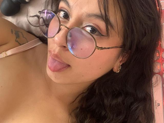MadyJackson with round glasses, sticking out her tongue, has dark wavy hair, a tattoo on her arm, and is wearing a pink bra, showing her bare shoulders.