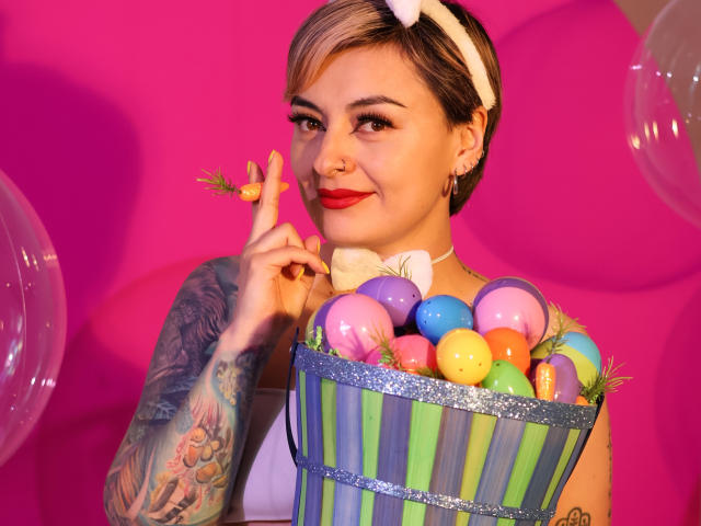 KatParadise con cabello rubio corto, un lazo blanco, lápiz labial rojo, sosteniendo huevos de Pascua en una cesta a rayas, brazo tatuado, fondo rosa, diadema blanca, sonriendo, sosteniendo una zanahoria..