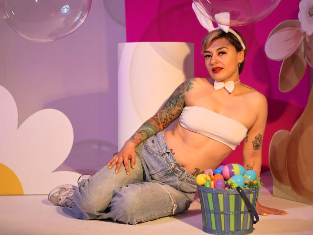 KatParadise, com cabelo curto, tatuada, vestindo um top branco sem mangas, calças jeans, orelhas de coelho, laço borboleta, segurando uma cesta de ovos de Páscoa, sentada, sorrindo, mostrando os seios, o bumbum e as tatuagens, em um cenário colorido e divertido..