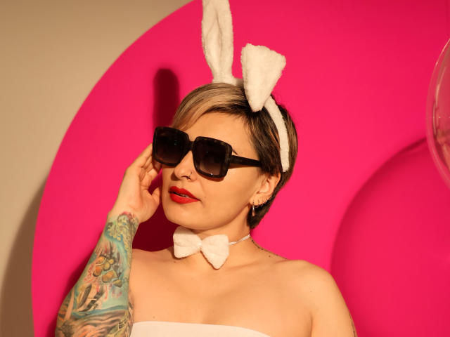 KatParadise, avec des cheveux blonds courts, des oreilles de lapin blanches, des lunettes de soleil noires, du rouge à lèvres rouge, un haut blanc sans bretelles et un bras tatoué, portant un nœud papillon blanc, sur un fond rose vif..