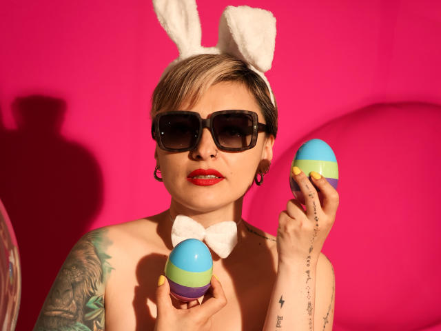 KatParadise with short blonde hair, white bunny ears, black sunglasses, red lipstick, tattoos, and a white bow, holding two colorful Easter eggs against a bright pink background.