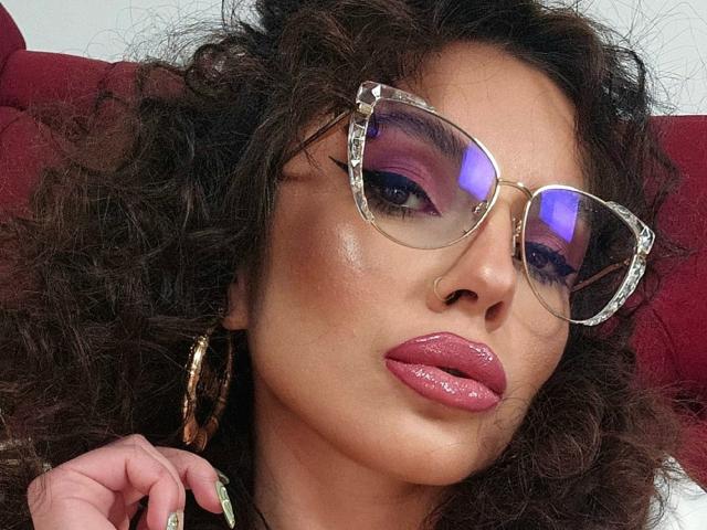 GoddessKaterina with curly brown hair, pink glossy lips, purple eyeshadow, reflective gold glasses, and gold hoop earrings, looking seductive with a hand near her face.