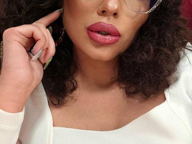 GoddessKaterina met glanzende, roze lippen, krullend donker haar, een bril, groene nagels, een witte top en een neusring, die haar oor aanraakt. Borsten niet zichtbaar..