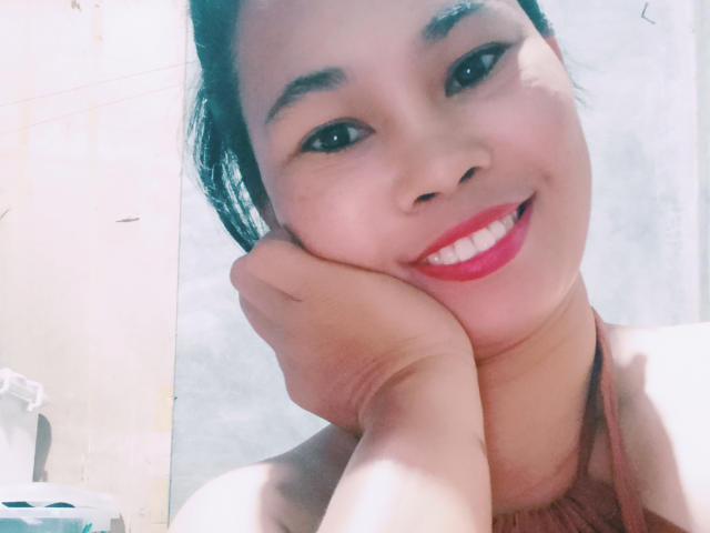 GiamayBeauty has a cute smile with red lipstick, black hair, and is resting her head on her hand. Her small tits are partially visible in a pink halter top.