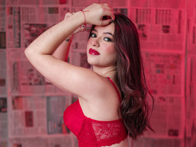 PaolaYKatty with red lace bra, raised arms, red lipstick, and long brown hair, looking over shoulder, red light on her. She's got big tits and a sexy smile.