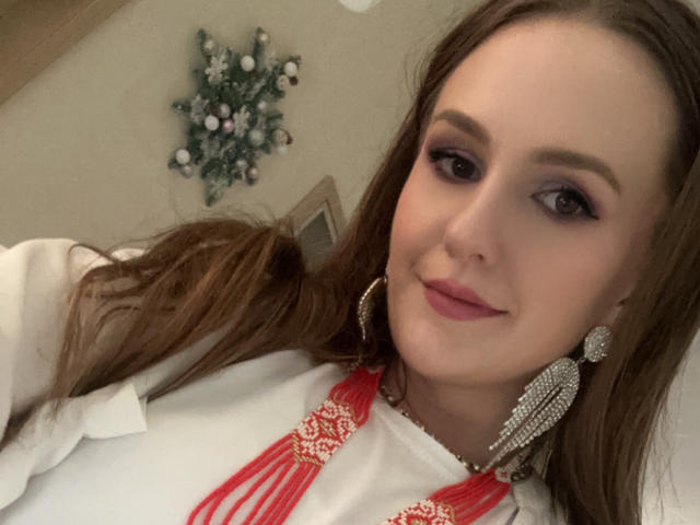 VanessaBeauty with long brown hair, purple eye makeup, pink lipstick, white top with red lace, and large diamond earrings, smiling softly. Her tits are partially visible.