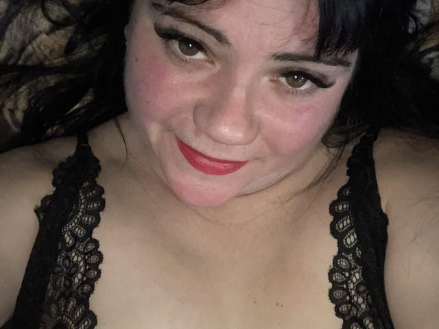 DebbieXRoy, with black lace top, smiling at camera, red lipstick, brown eyes, black hair, fair skin, showing her tits, lying down, slight blush, cute, sexy, confident.