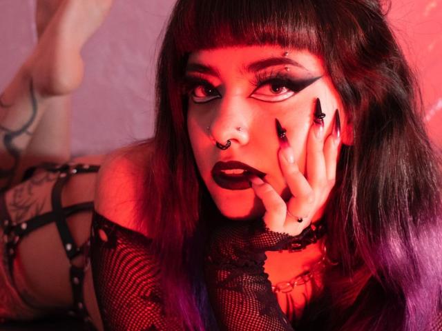 LitenkittenOne has dark lipstick, dramatic eyeliner, and a nose ring. She's in black lace, with purple and black hair, showing her hand and leg tattoos. Red lighting.