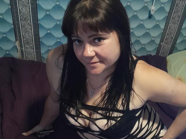 DebbieXRoy with black strappy lingerie, exposing her tits, sitting on a bed with purple and green pillows, dark brown hair, looking up, slight smile, blue quilted wall behind.