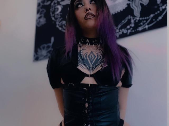 LitenkittenOne with purple and black hair, black goth makeup, and tattoos, wearing a black leather corset and choker, looking up with hands behind back, in a dark room.