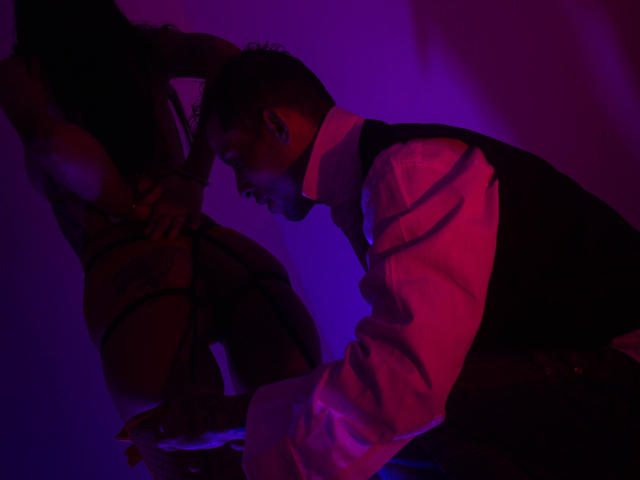 BenedettoMichelRuby with dark skin and short curly hair, wearing a vest and shirt, leans forward, eyes on BenedettoMichelRuby's bare ass, lit by purple and pink lights.