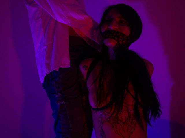BenedettoMichelRuby with dark hair, gagged, topless, wearing black mesh, bent over, white shirt draped, purple and pink lighting, exposing her tits and tattoo on her right arm.