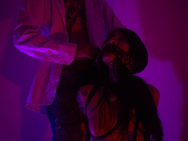 BenedettoMichelRuby with black hair, gagged with a checkered mask, wearing a hat, next to BenedettoMichelRuby in a white coat, purple and pink lighting, tight grip on neck.